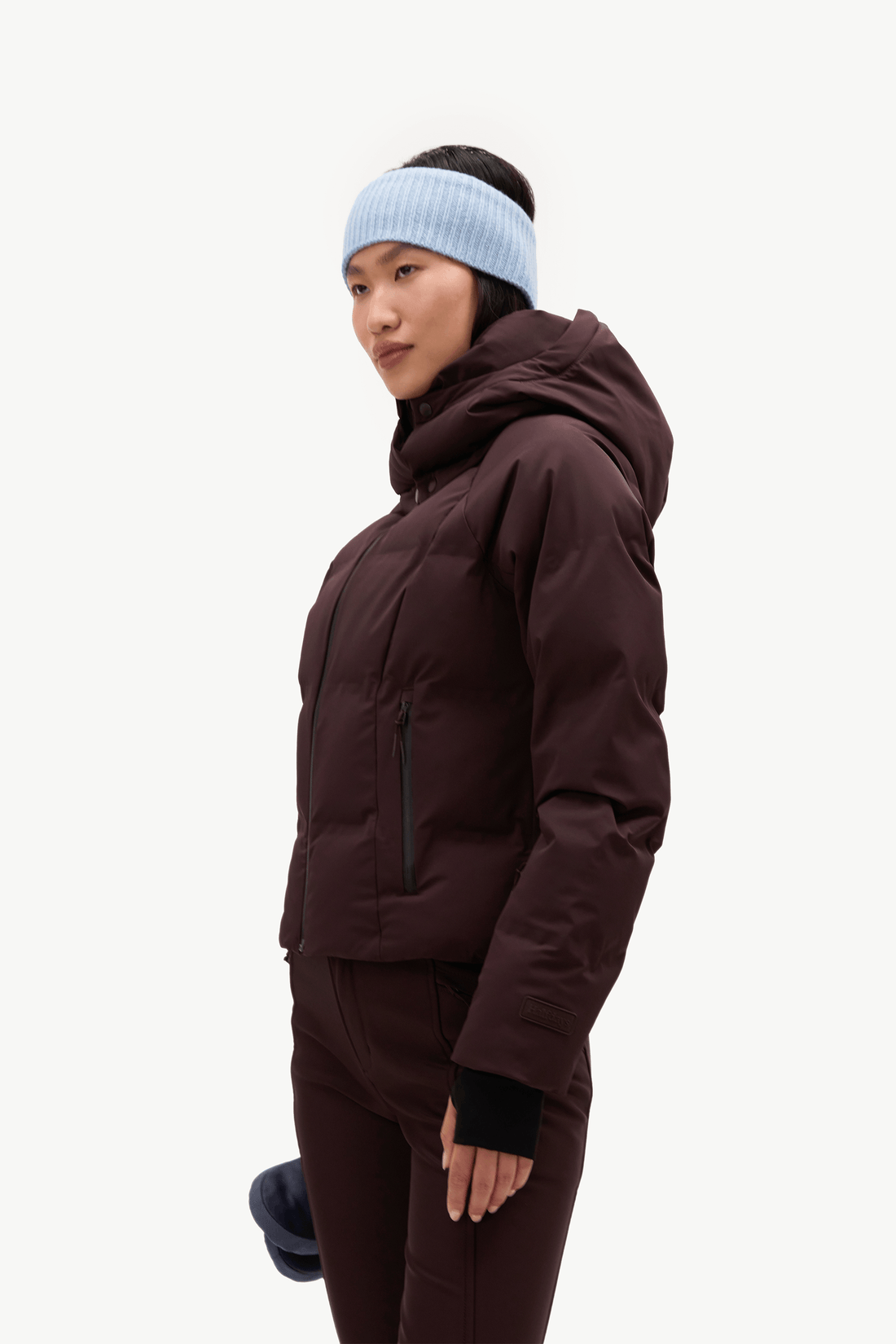 A woman wearing a dark brown hooded puffer jacket, matching dark brown pants, a light blue knit beanie, and a black beanie, photographed against a solid off-white background.