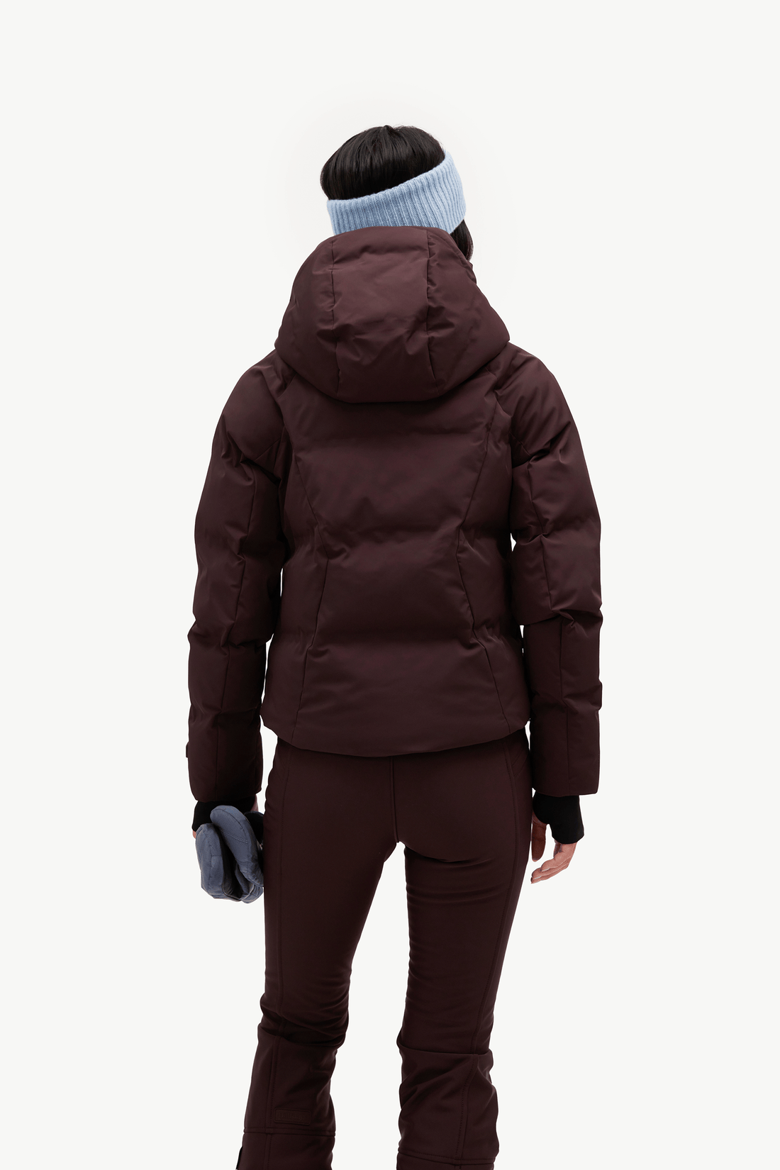 A model from behind wearing a dark burgundy hooded puffer jacket, matching dark pants, a light blue knit beanie against a clean white background.