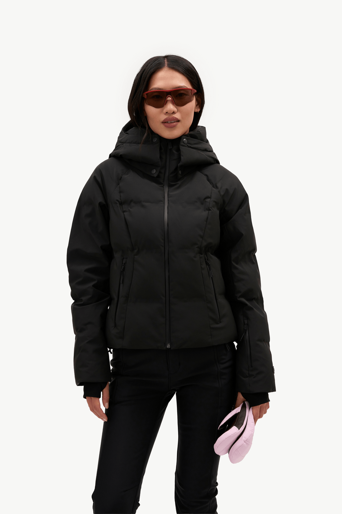 A model wearing a black hooded puffer jacket, black pants, and sunglasses, holding a light pink accessory against a plain white background.