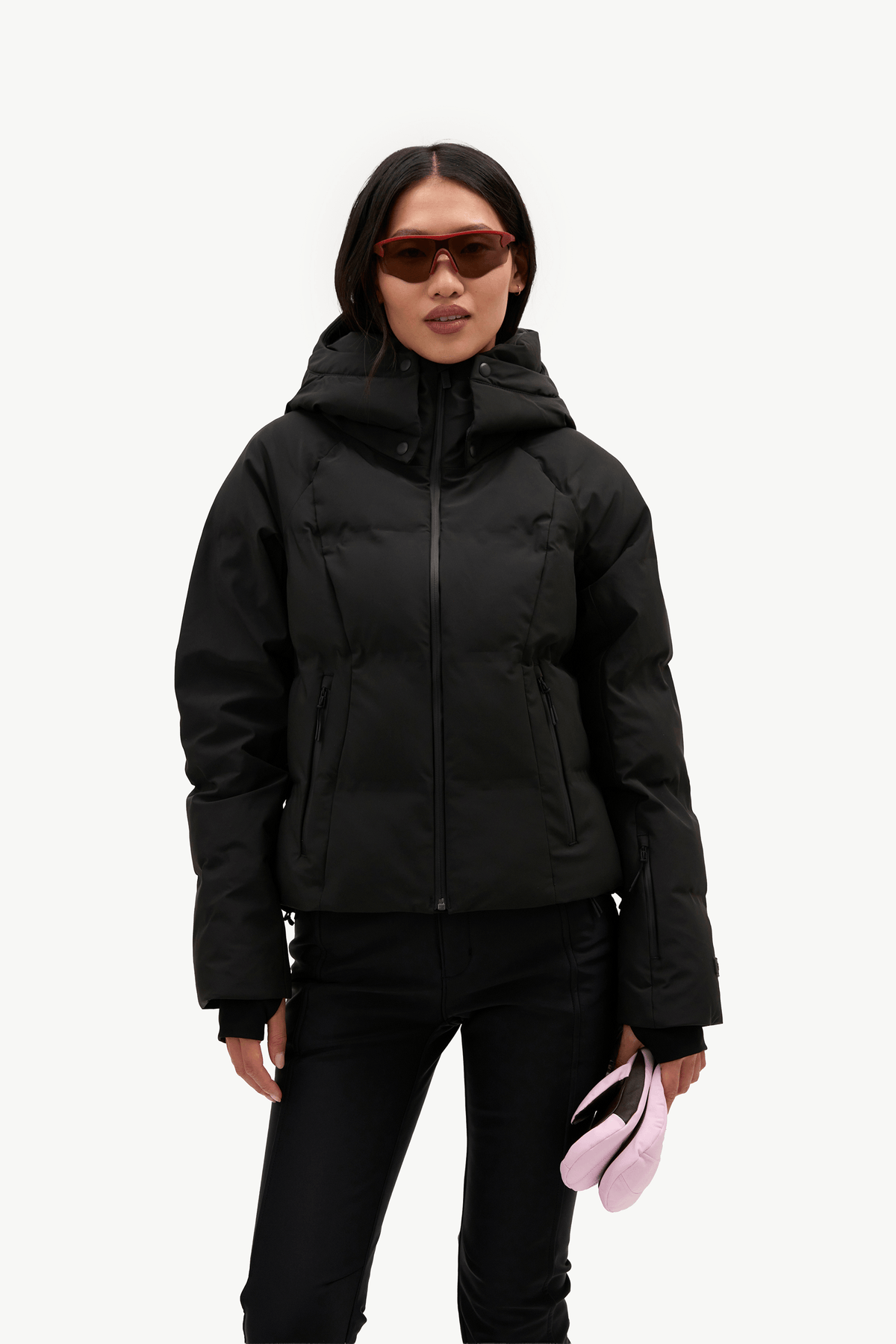 A model wearing a black hooded puffer jacket, black pants, and sunglasses, holding a light pink accessory against a plain white background.