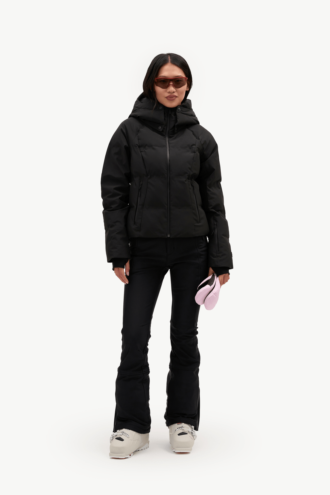A model stands against a plain white studio background wearing a black hooded puffer jacket, black pants, and white sneakers, while holding a small pink item in one hand.