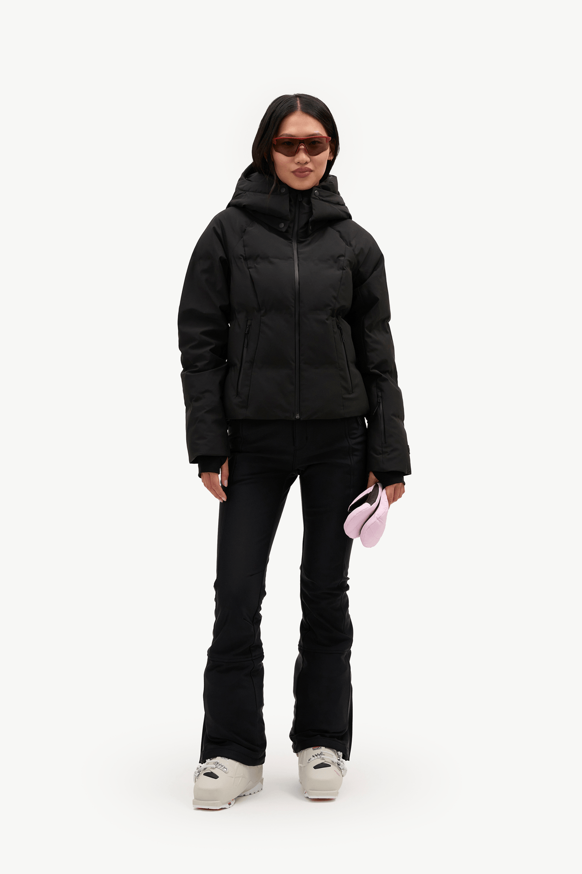 A model stands against a plain white studio background wearing a black hooded puffer jacket, black pants, and white sneakers, while holding a small pink item in one hand.