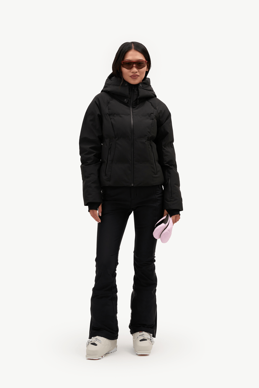 A model stands against a plain white studio background wearing a black hooded puffer jacket, black pants, and white sneakers, while holding a small pink item in one hand.