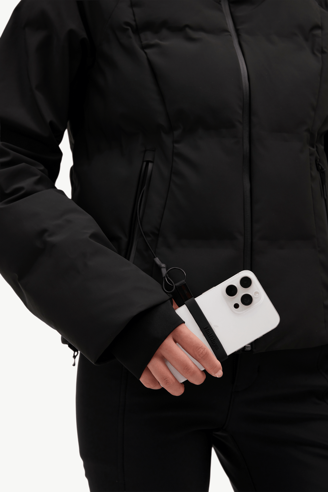 A close-up of a person wearing a black puffer jacket and black pants, holding a white smartphone with a black wrist strap accessory attached near the cuff.