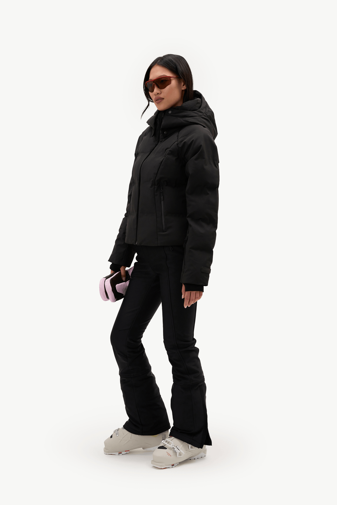 A woman modeling a black hooded puffer jacket, black ski pants, and light gray sneakers against a plain white studio background.