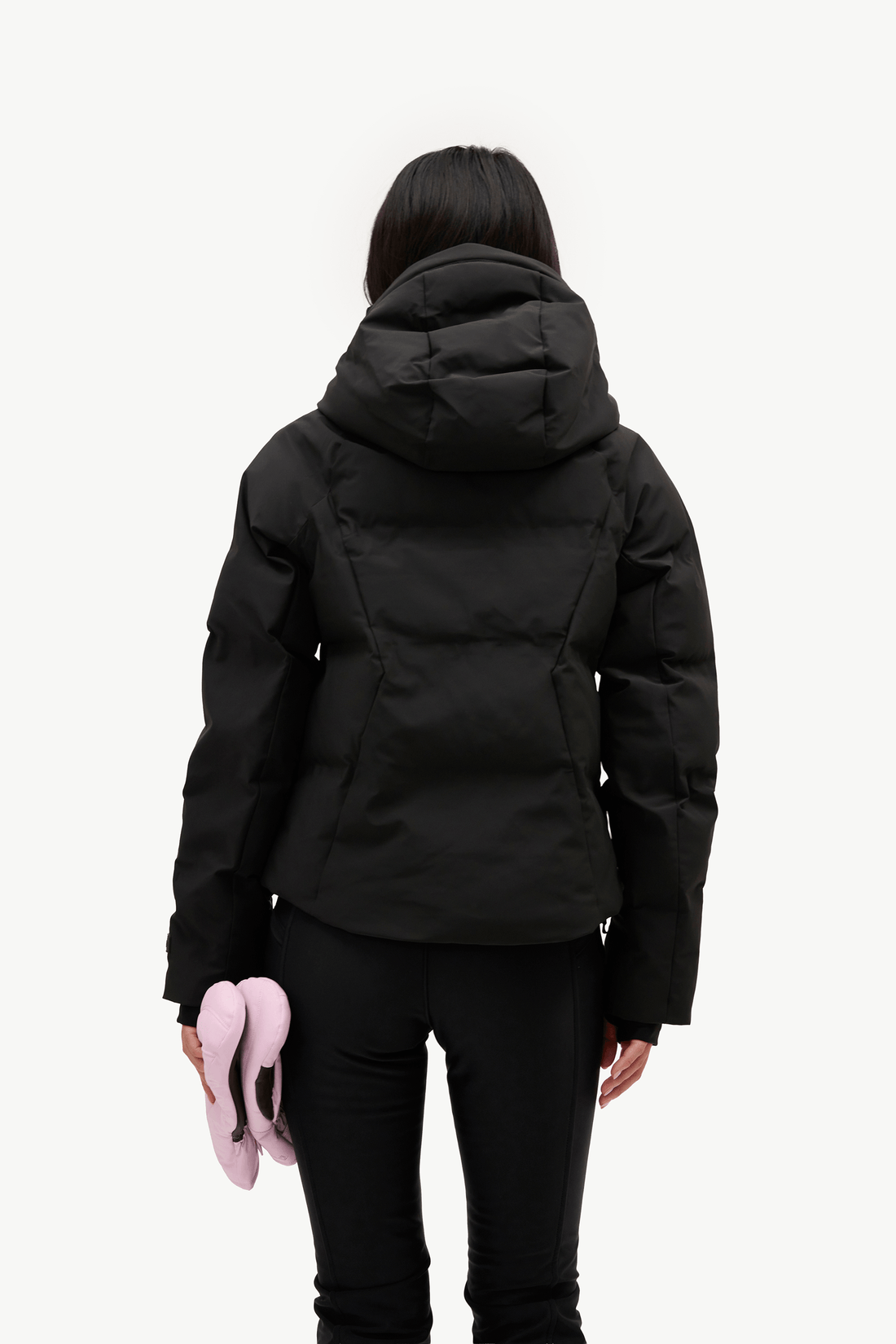 A woman viewed from behind wearing a black puffer jacket and black pants, holding a pair of light pink slippers against a plain white studio background.