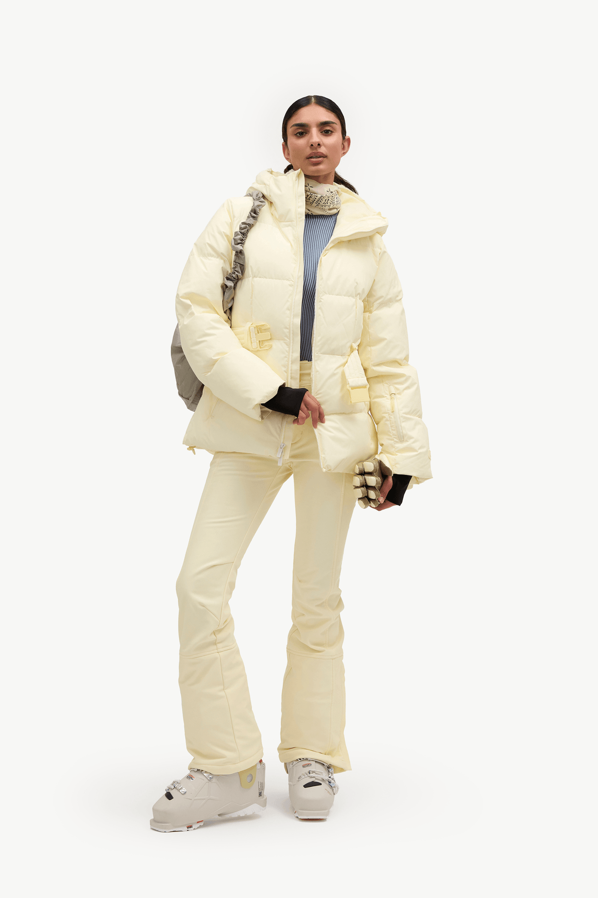 A model stands against a plain white studio background wearing a pale cream puffer jacket and matching ski pants, with a blue top underneath and white ski boots.