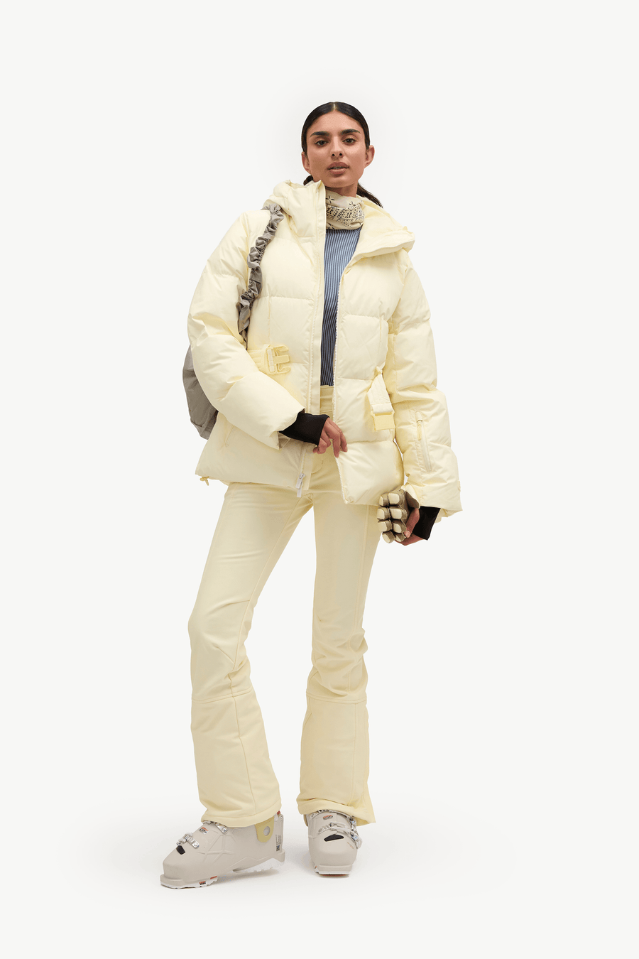 A model stands against a plain white studio background wearing a pale cream puffer jacket and matching ski pants, with a blue top underneath and white ski boots.