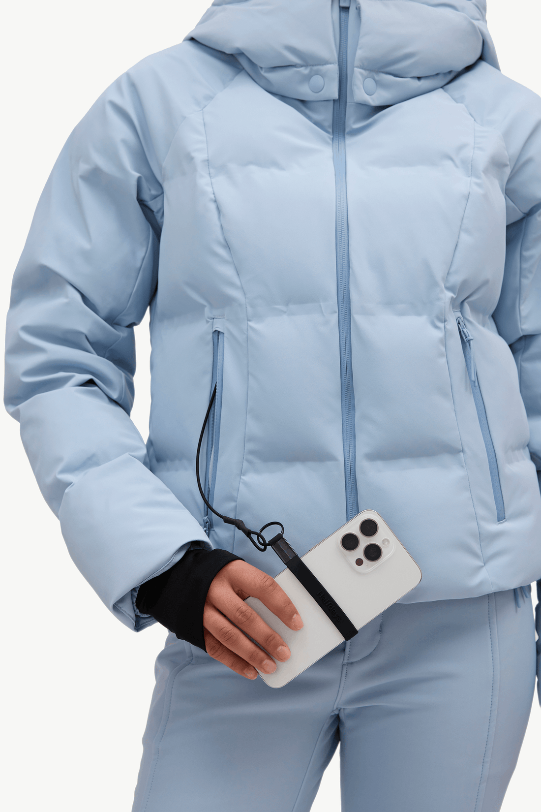 A person wearing a light blue hooded puffer jacket and matching light blue pants, holding a white smartphone with a black wrist strap accessory attached near the pocket.