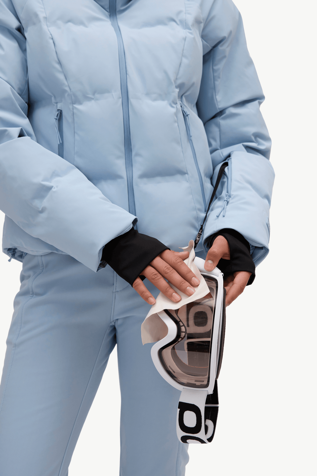 A woman in a light blue puffer jacket and matching flared ski pants, holding goggles against a plain white studio background.