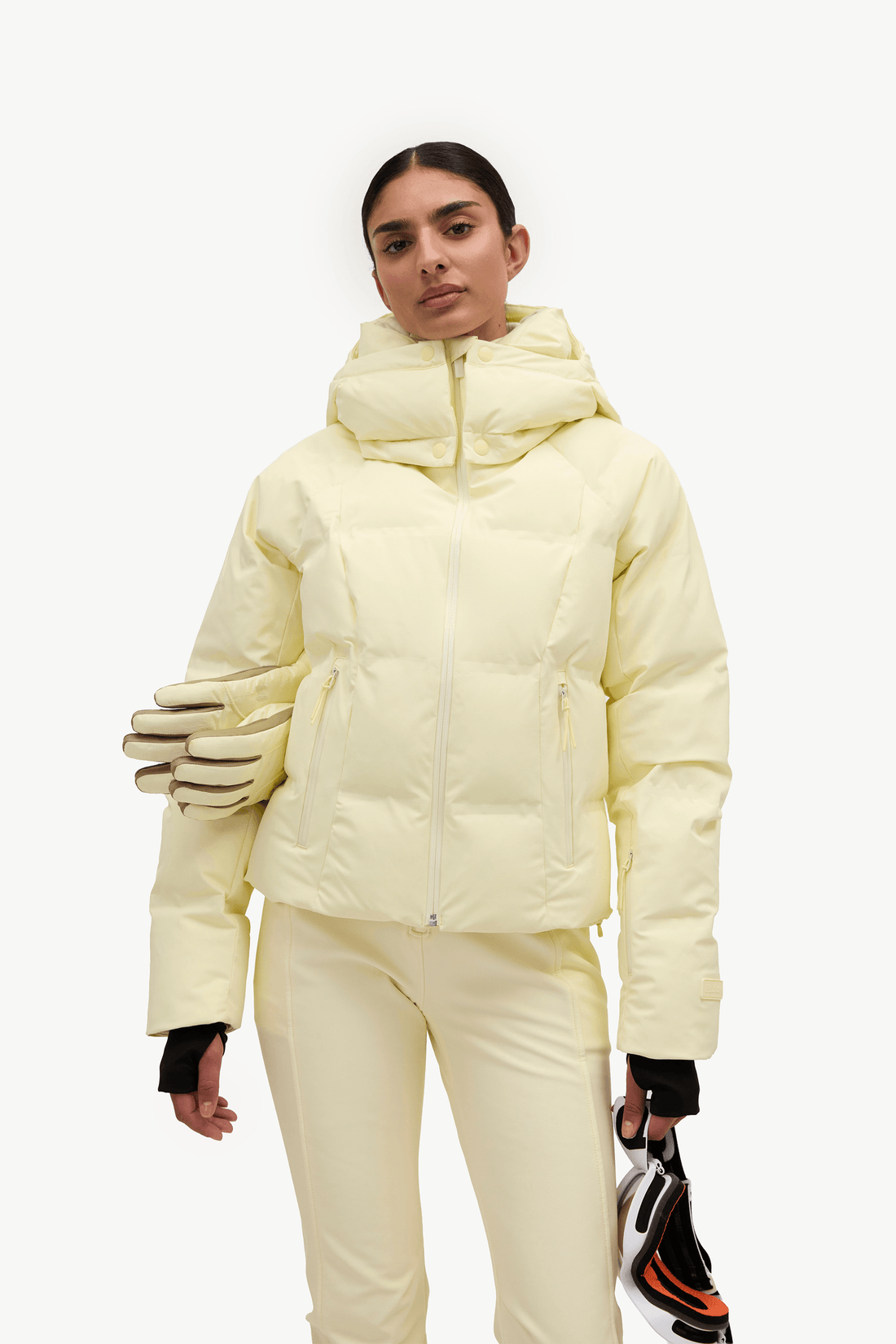 A model wearing a pale yellow puffer jacket and matching ski pants, holding a pair of gloves and a ski boot against a plain white studio background.