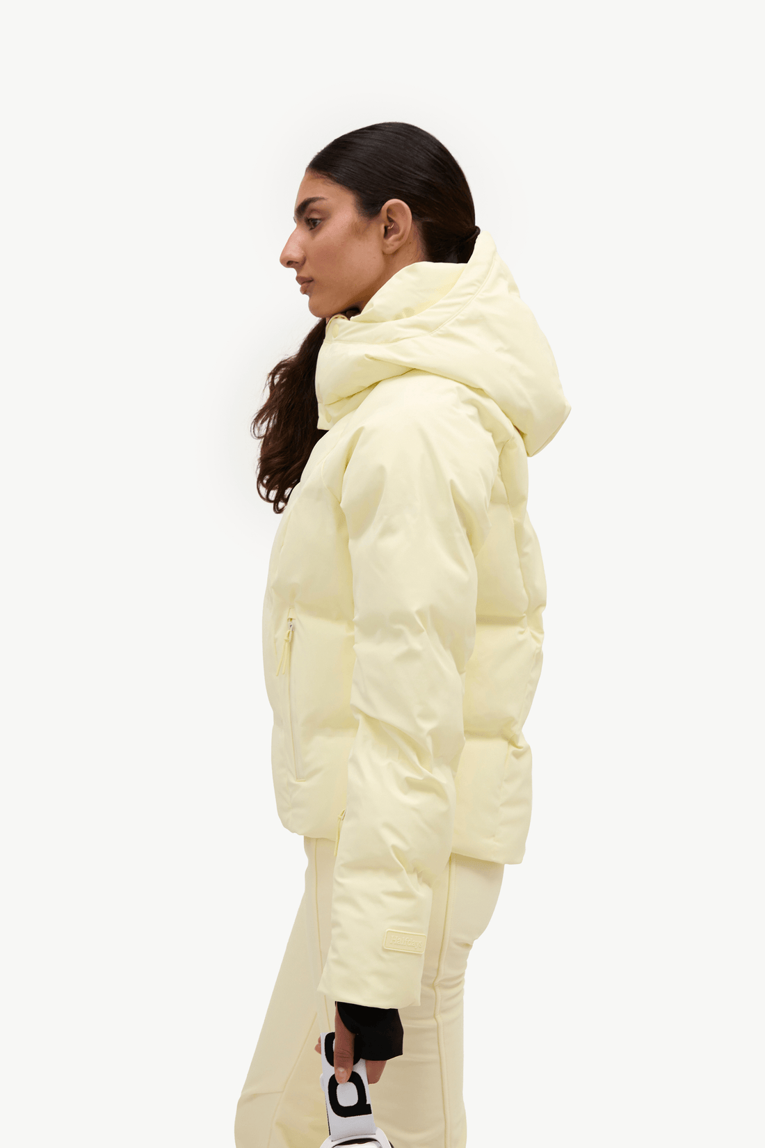 A woman in profile wearing a pale pastel yellow puffer jacket and matching light pants against a clean white studio background.