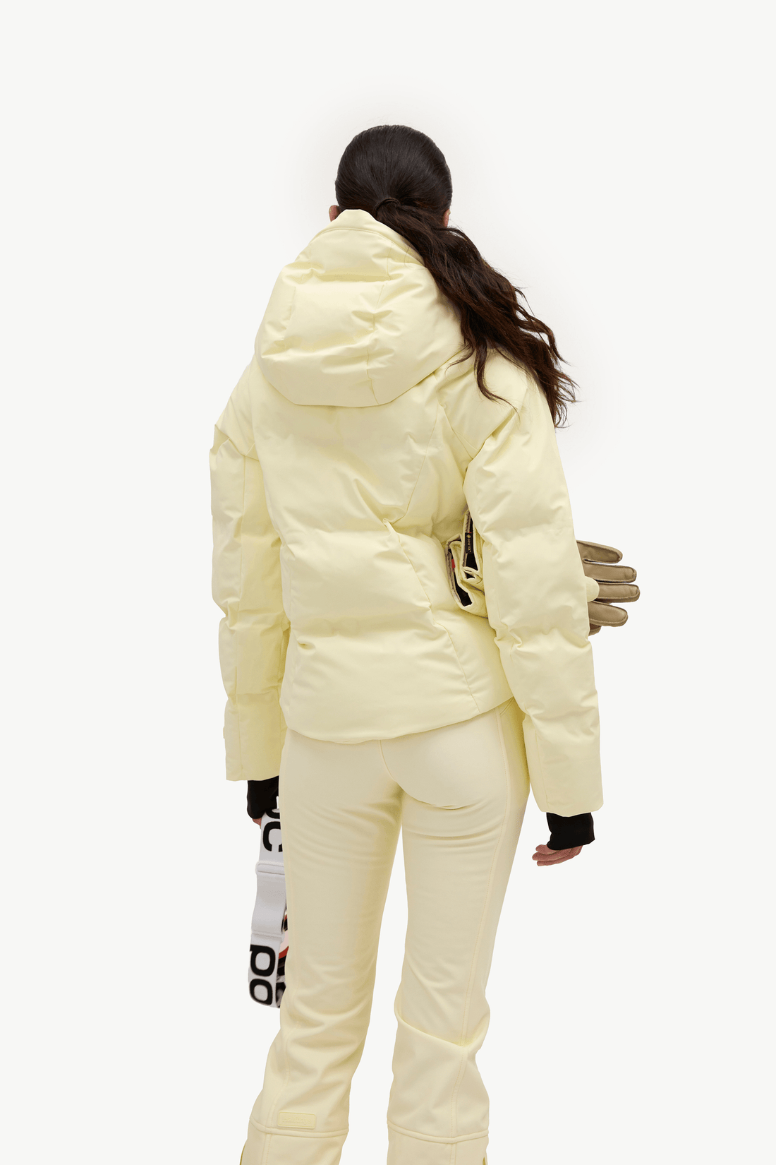 A woman from behind wearing a pale pastel yellow puffer jacket and matching light pants against a clean white studio background.