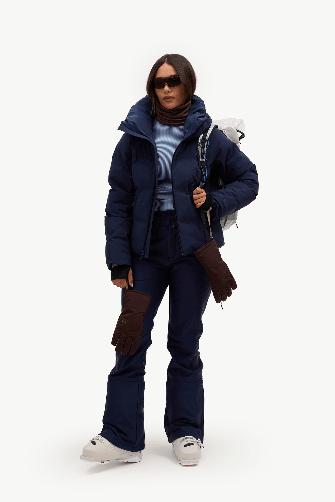 A model wearing a dark navy puffer jacket, matching navy pants, a light blue ribbed turtleneck, brown gloves, and white sneakers, standing against a plain white studio background.