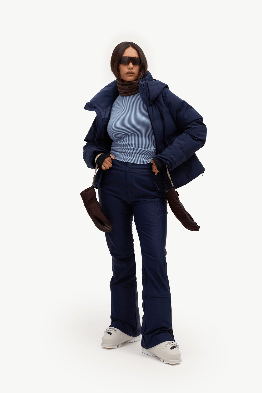 A model wearing a dark navy puffer jacket, matching navy pants, a light blue ribbed turtleneck, brown gloves, and white sneakers, standing against a plain white studio background.