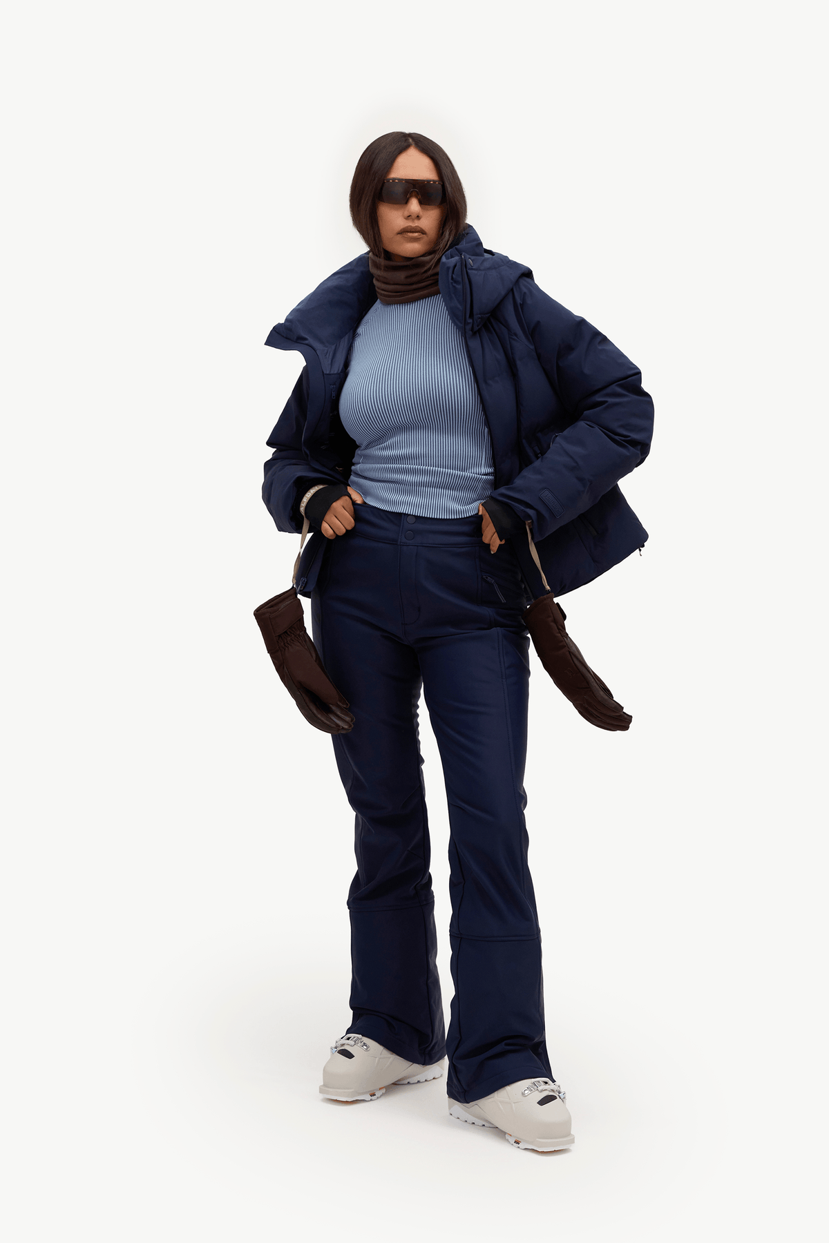 A model wearing a dark navy puffer jacket, matching navy pants, a light blue ribbed turtleneck, brown gloves, and white sneakers, standing against a plain white studio background.