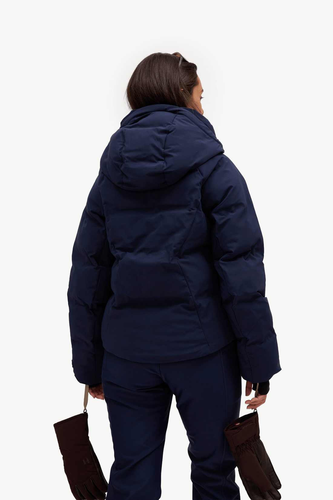 A model wearing a dark navy puffer jacket, matching navy pants, a light blue ribbed turtleneck, brown gloves, standing against a plain white studio background.