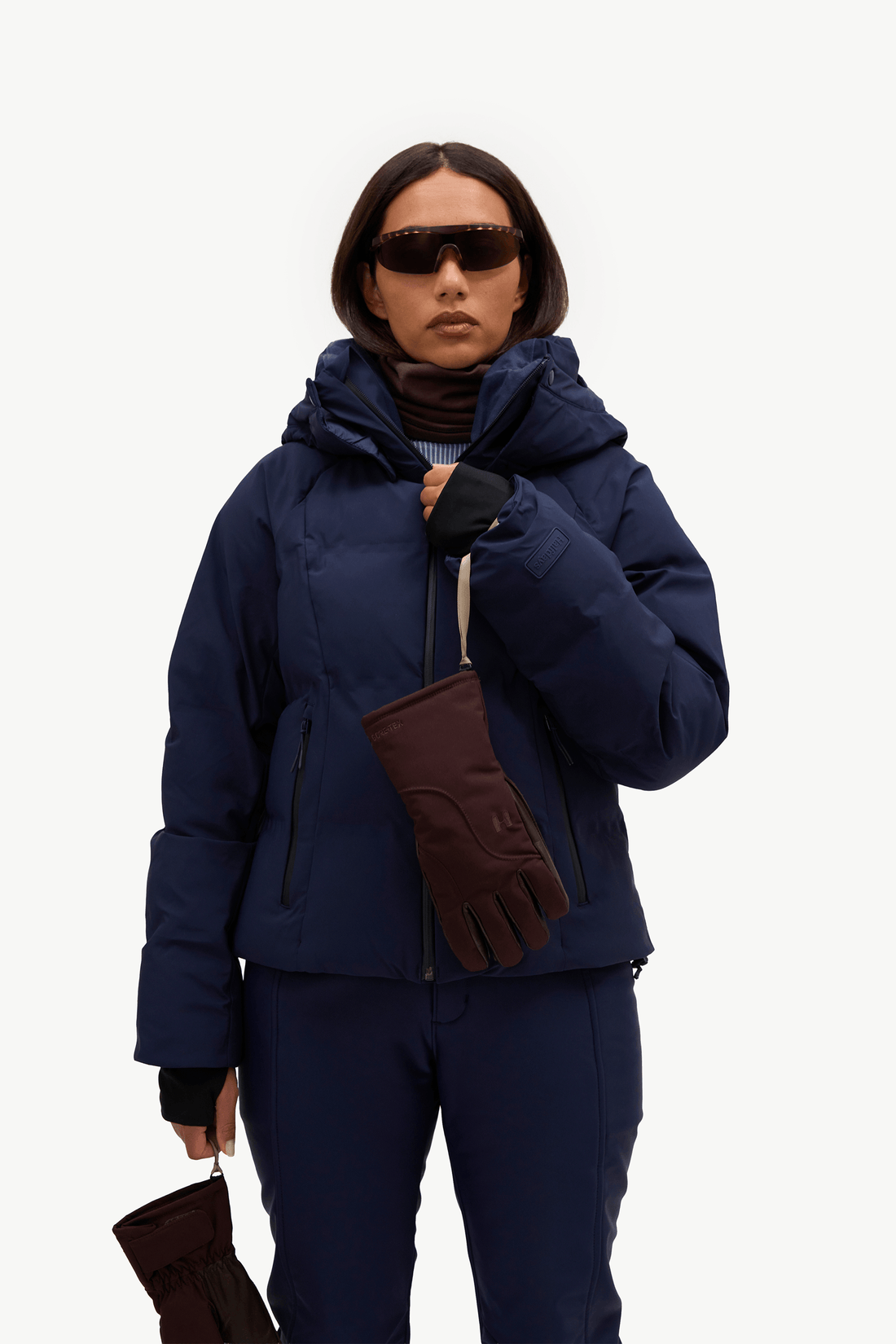 A model wearing a dark navy puffer jacket, matching navy pants, a light blue ribbed turtleneck, brown gloves, standing against a plain white studio background.