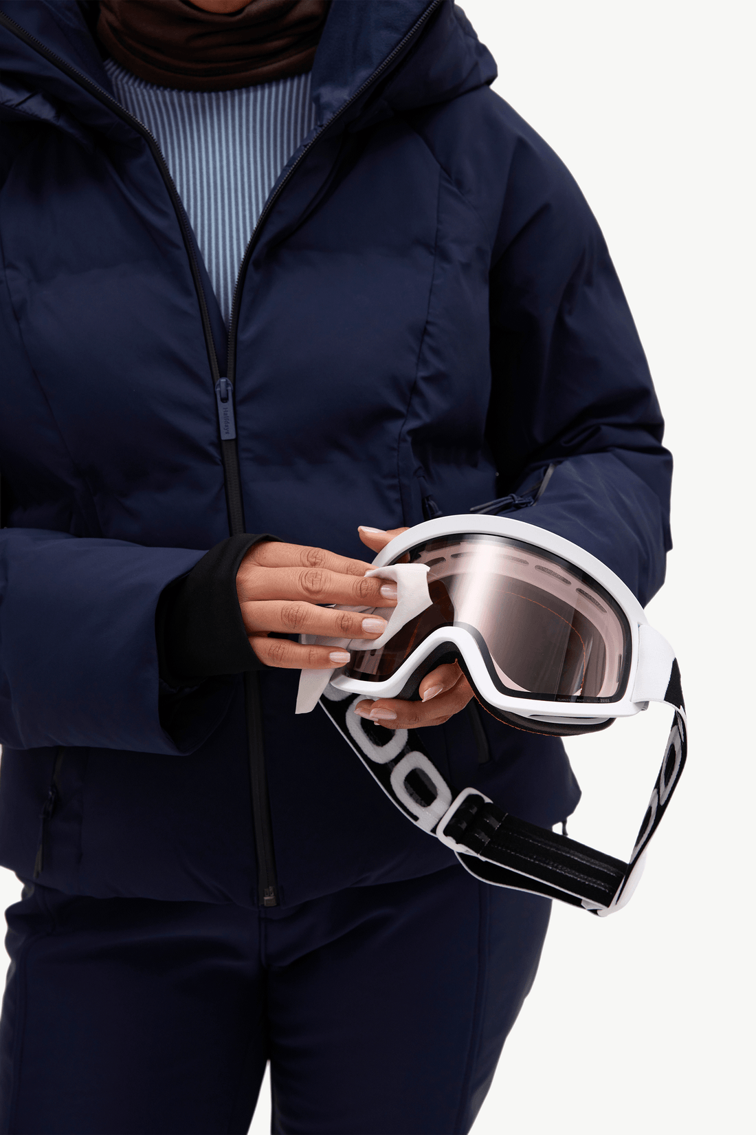 A model wearing a dark navy puffer jacket, matching navy pants, a light blue ribbed turtleneck,wiping goggles standing against a plain white studio background.