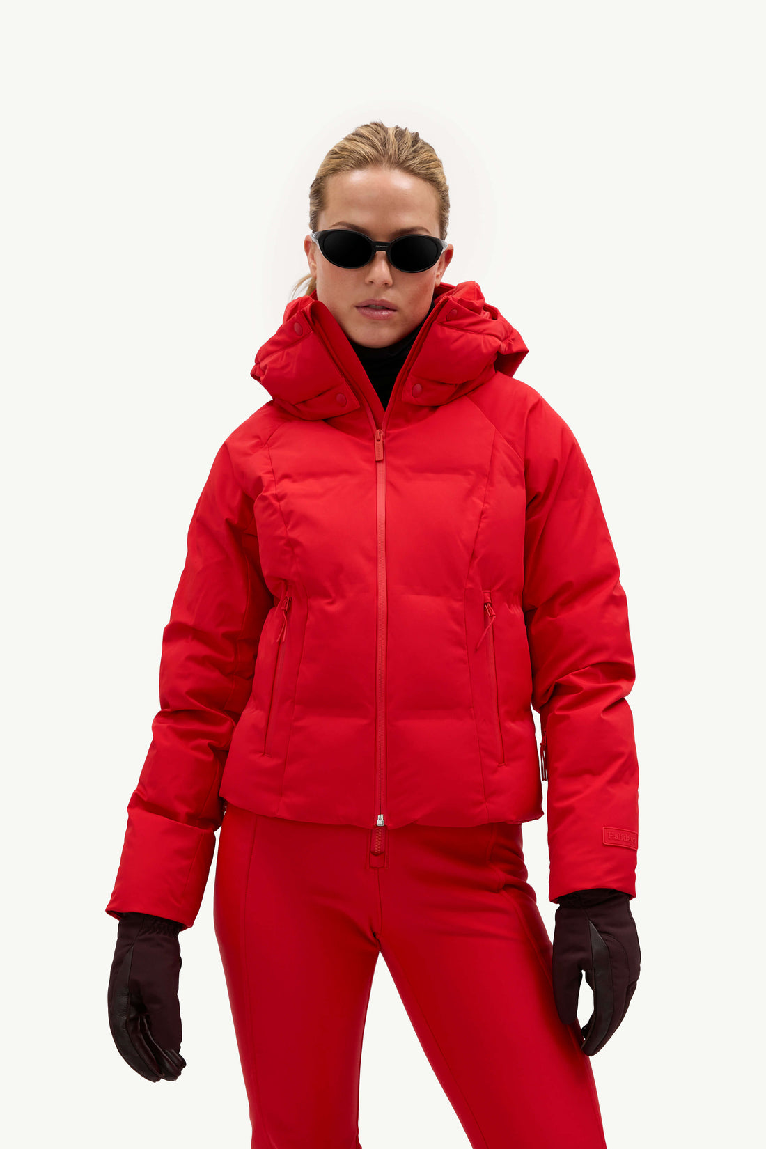 A model wearing a bright red ski jacket and matching red ski pants, styled with black gloves, black sunglasses, and white ski boots against a plain white studio background.