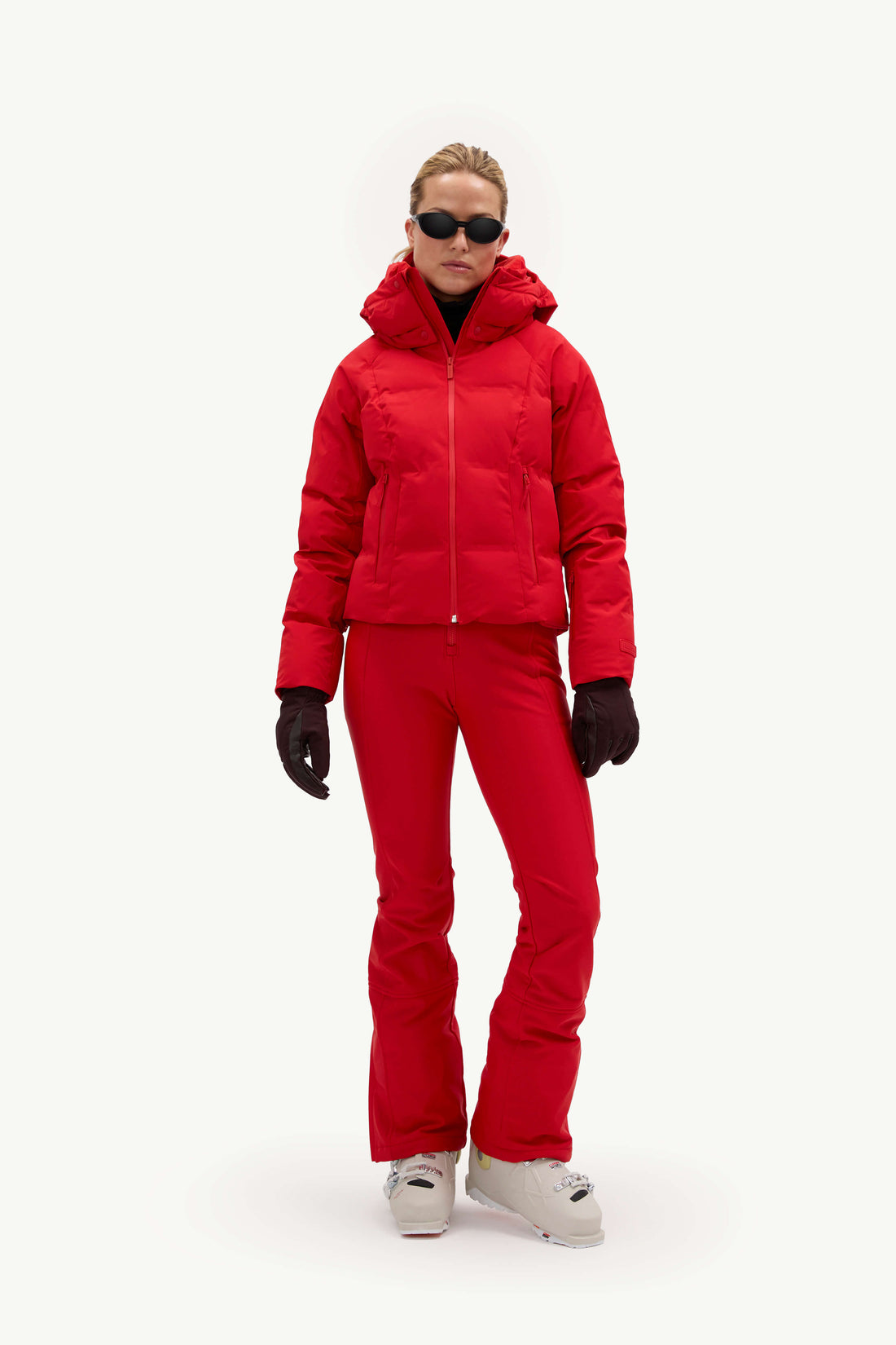 A model wearing a bright red ski jacket and matching red ski pants, styled with black gloves, black sunglasses, and white ski boots against a plain white studio background.