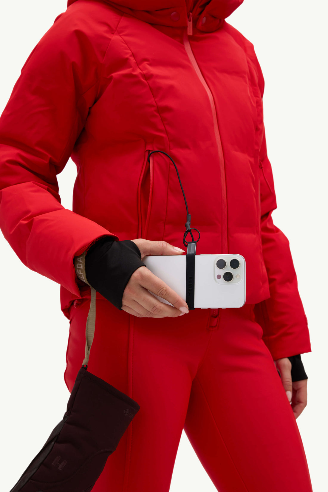 A close-up of a person wearing a bright red hooded puffer jacket and matching red pants, holding a white smartphone with a black strap accessory attached near the waist.