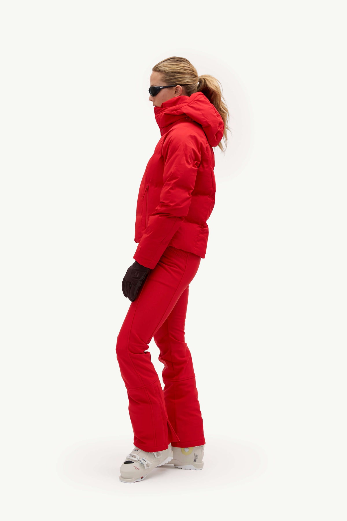 A model wearing a bright red ski jacket and matching red ski pants, styled with black gloves, black sunglasses, and white ski boots against a plain white studio background.