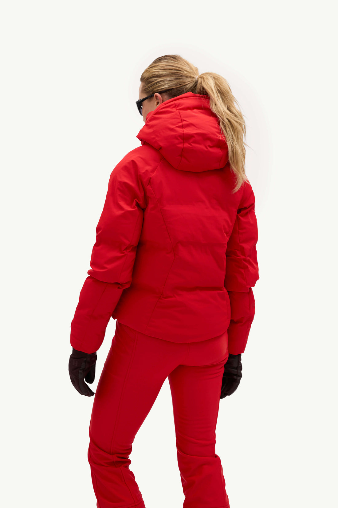 A woman wearing a bright red puffer jacket and matching red ski pants, shown from the back against a plain white studio background.
