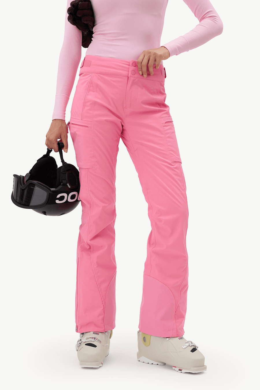 A model wearing bright pink ski pants, a light pink long-sleeve top, and beige ski boots, holding a black O'Neill helmet against a plain white studio background.