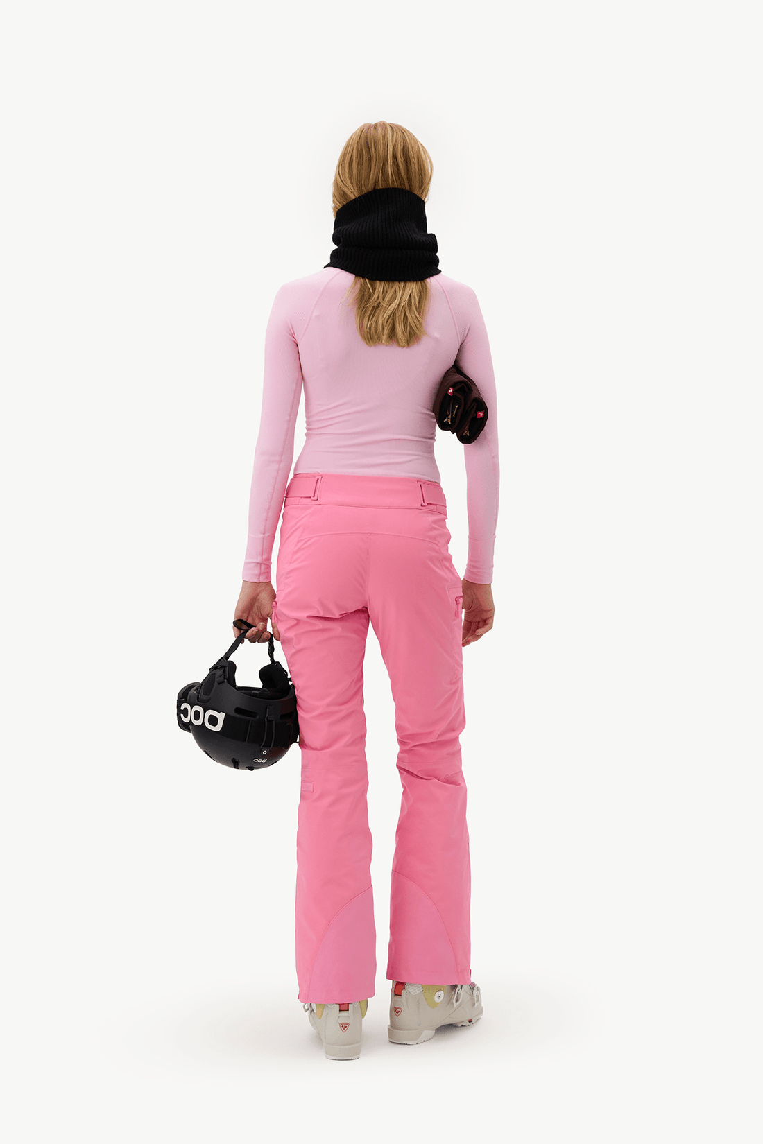 A woman viewed from behind wearing a light pink long-sleeve top and bright pink ski pants, with a black neck gaiter and carrying black ski gear against a plain white studio background.