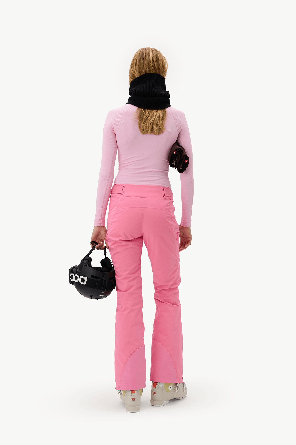 A woman viewed from behind wearing a light pink long-sleeve top and bright pink ski pants, with a black neck gaiter and carrying black ski gear against a plain white studio background.