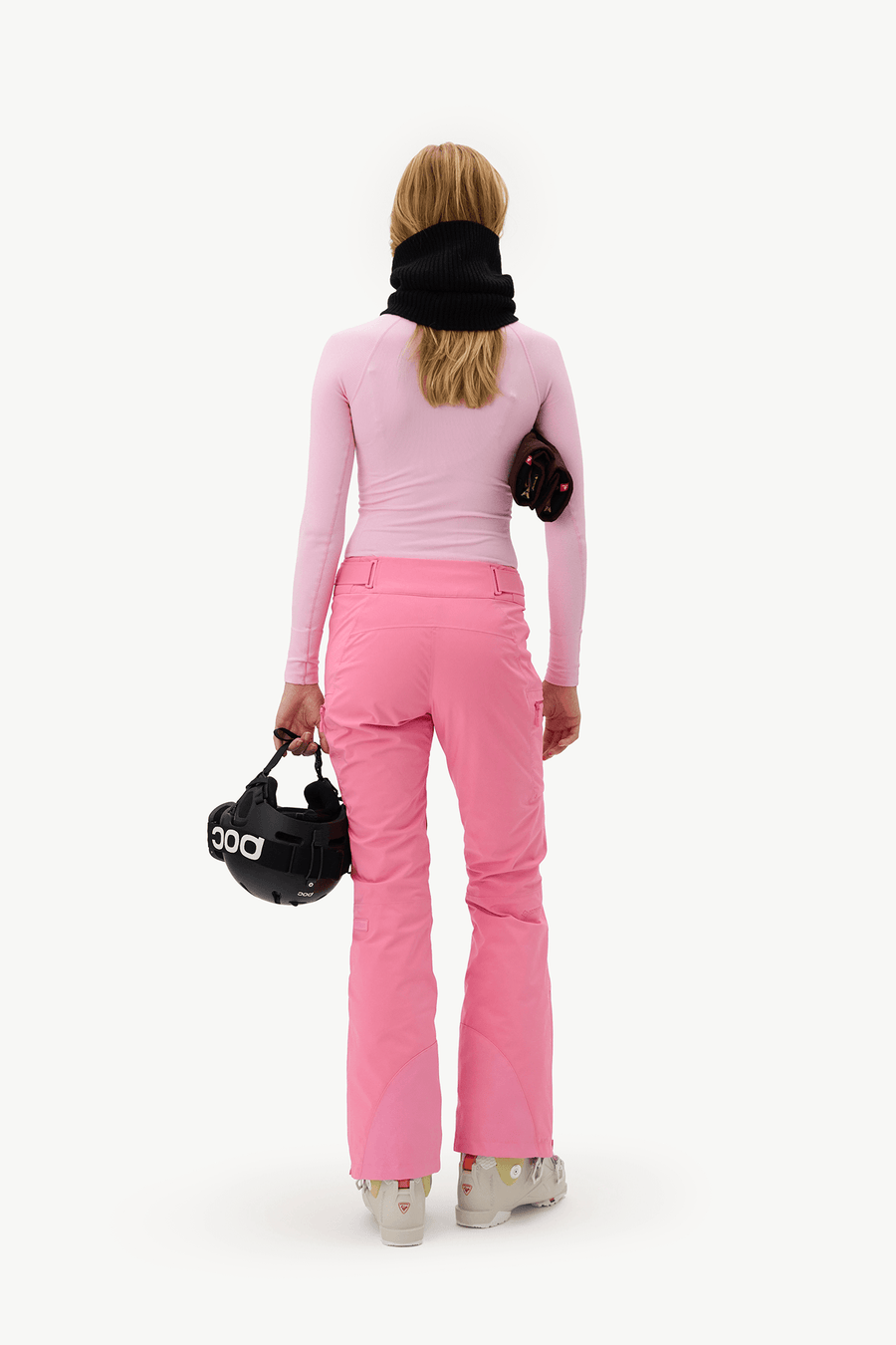 A woman viewed from behind wearing a light pink long-sleeve top and bright pink ski pants, with a black neck gaiter and carrying black ski gear against a plain white studio background.