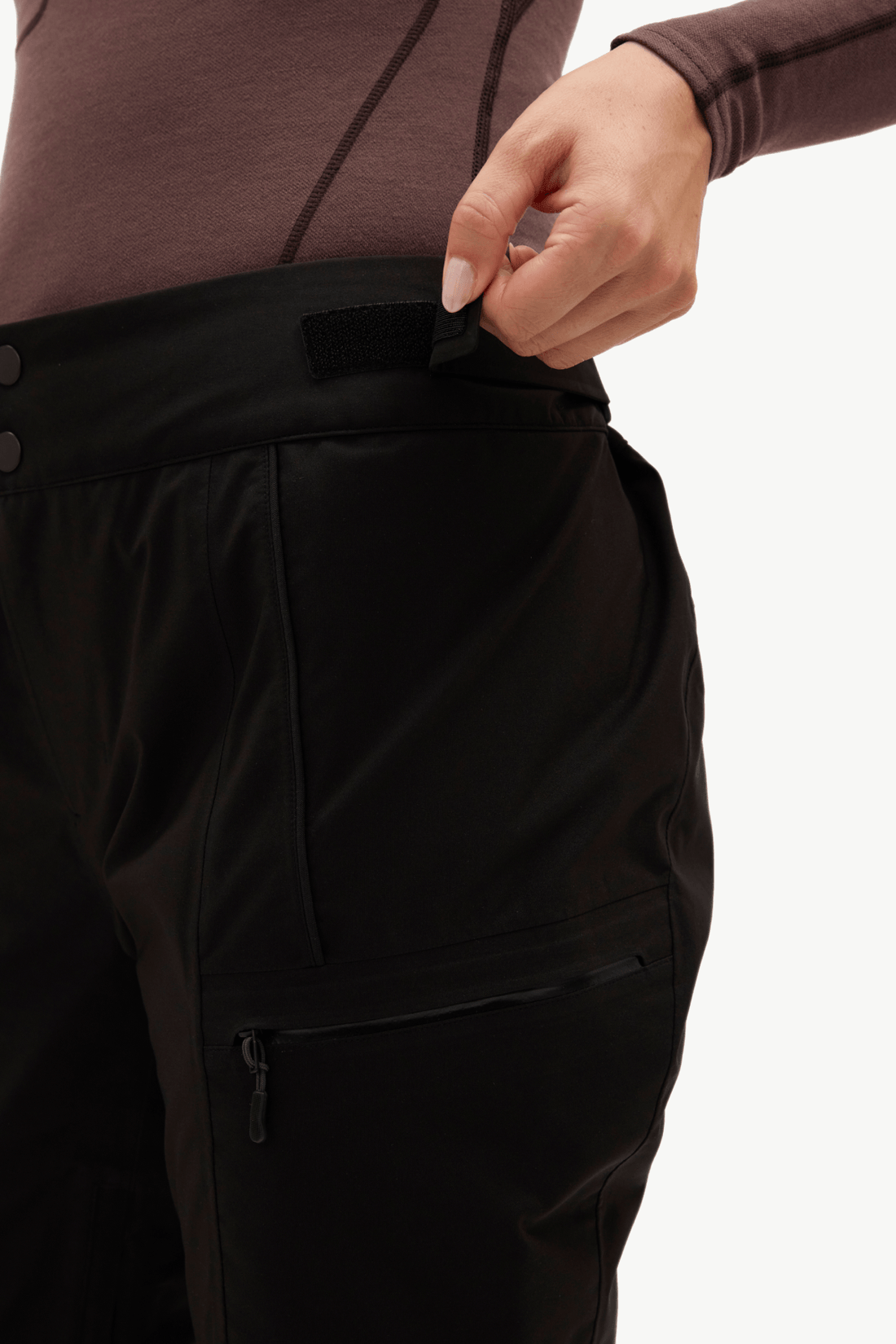 Close-up of a person wearing black technical pants, pulling a black hook-and-loop closure tab on the waistband with one hand. The person is also wearing a brown long-sleeve athletic top against a clean white background.