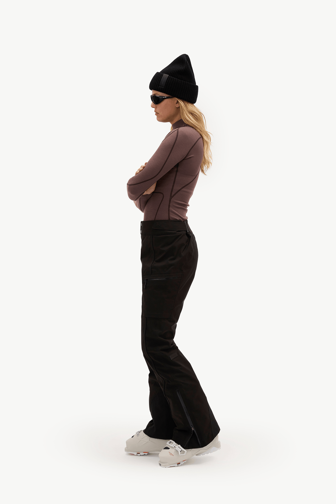 A woman stands in profile wearing a black knit beanie, black ski pants, a fitted taupe-brown long-sleeve top, black sunglasses, and white sneakers against a plain white studio background.
