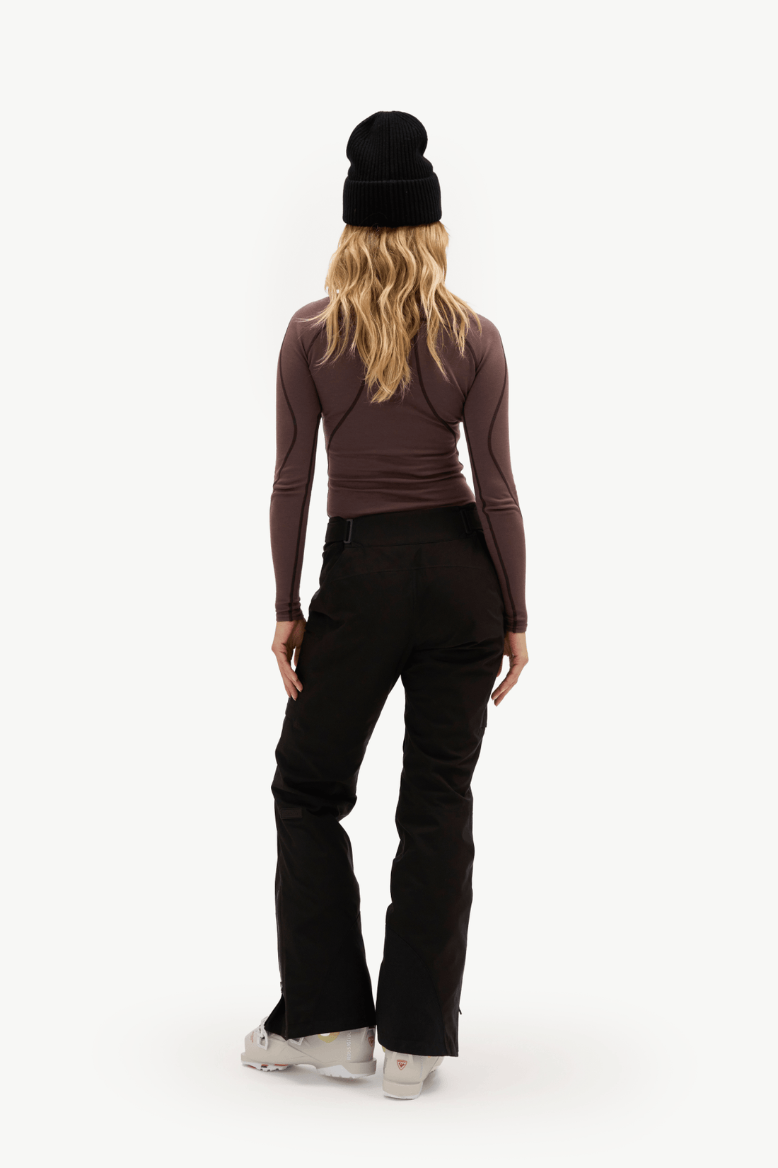 A woman is shown from behind wearing a black knit beanie, a dark brown long-sleeve top, black flared pants, and white sneakers against a plain white studio background.