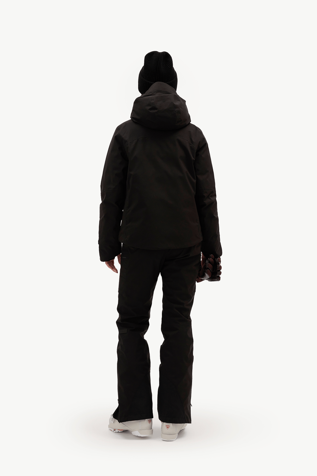 A person shown from the back wearing a black hooded puffer jacket, black pants, a black beanie, and white sneakers against a plain white studio background.