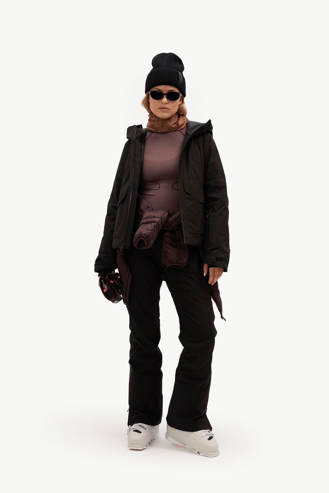 A model stands against a plain white studio background wearing a full ski outfit: a black beanie, black sunglasses, a black jacket over a dark brown top, black ski pants, and white ski boots.