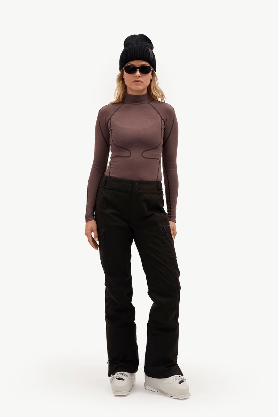 A model wearing a fitted long-sleeve athletic top in dark brown, black snow pants, a black beanie, sunglasses, and white ski boots, standing against a plain white studio background.
