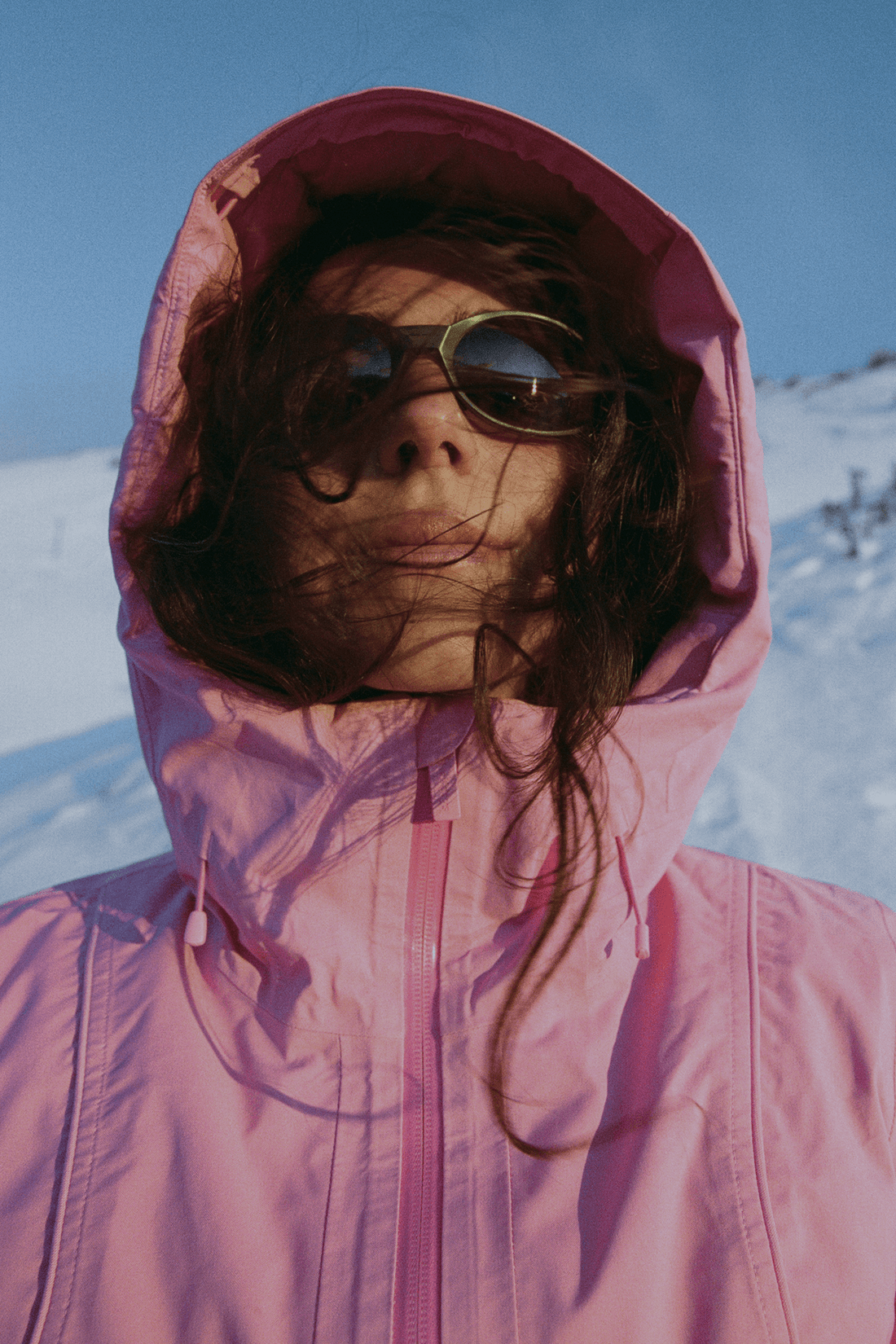 A person wearing a pink hooded jacket and sunglasses, photographed outdoors against a snowy landscape and blue sky.