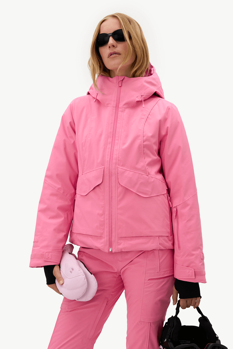 A woman modeling a bright pink ski jacket and matching pink pants against a clean white background, holding a pink helmet and a black bag.