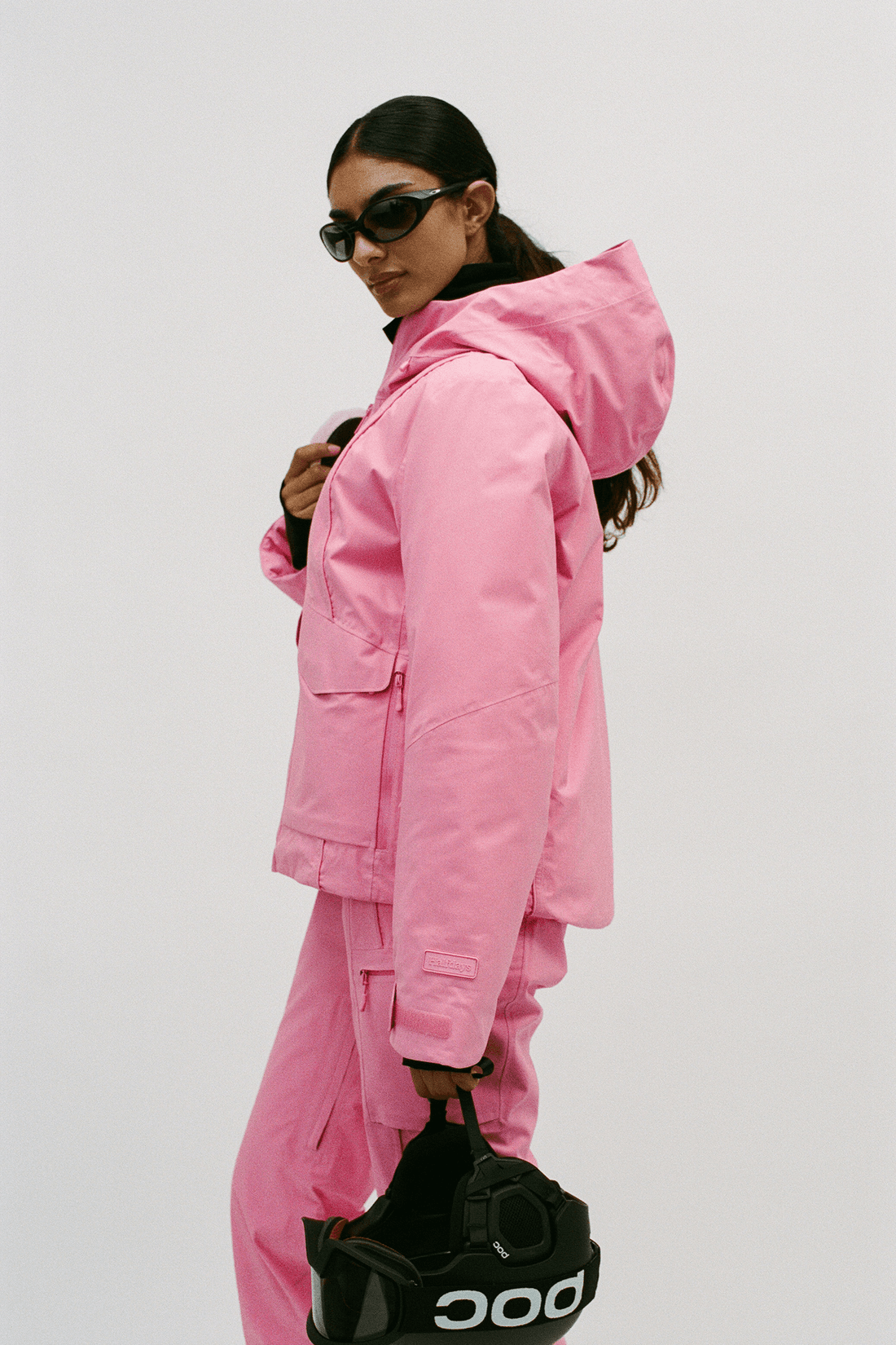 A model wearing a matching pink hooded jacket and pants set, styled with black sunglasses and holding a black POC bag against a plain light gray background.