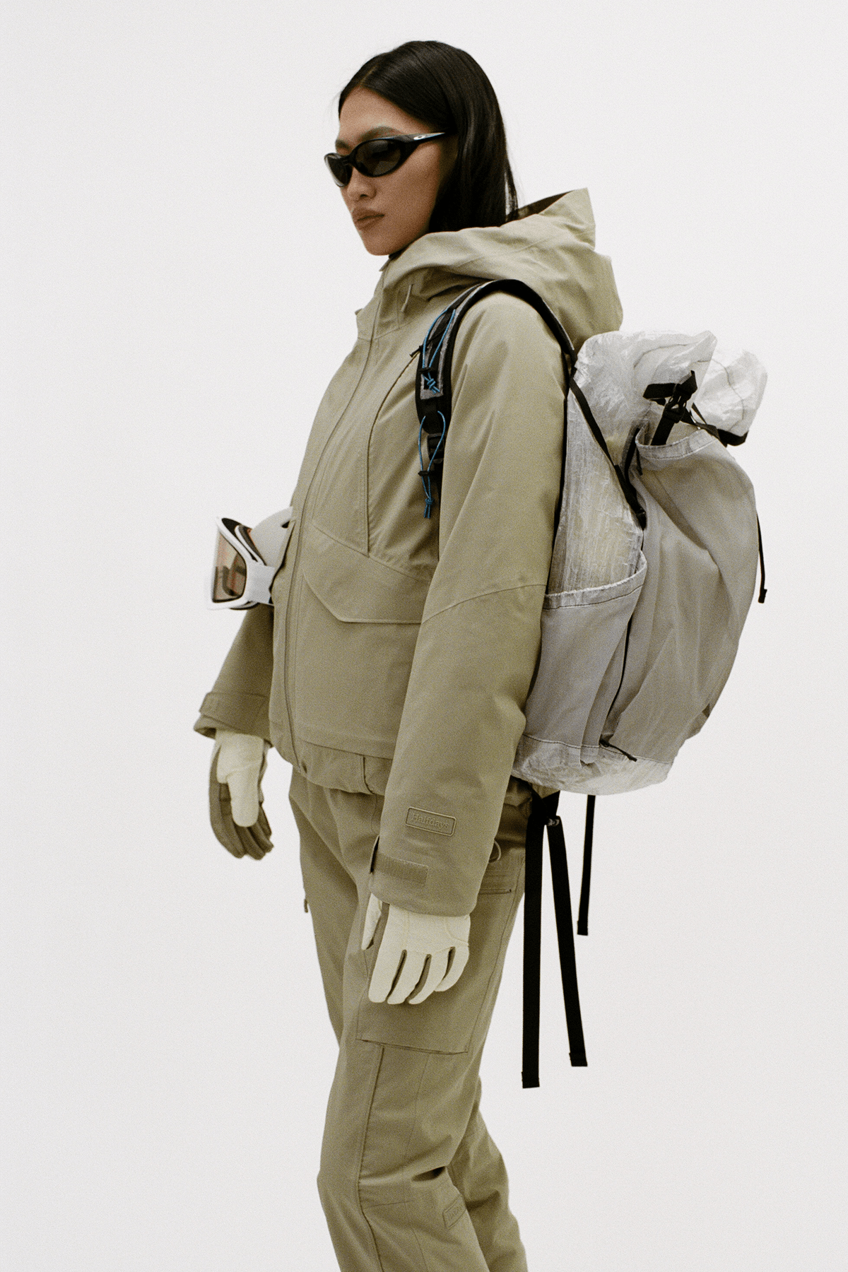A model wearing a beige technical outdoor jacket and matching pants, accessorized with a large translucent gray backpack, white gloves, and black sunglasses, photographed against a plain light gray studio background.