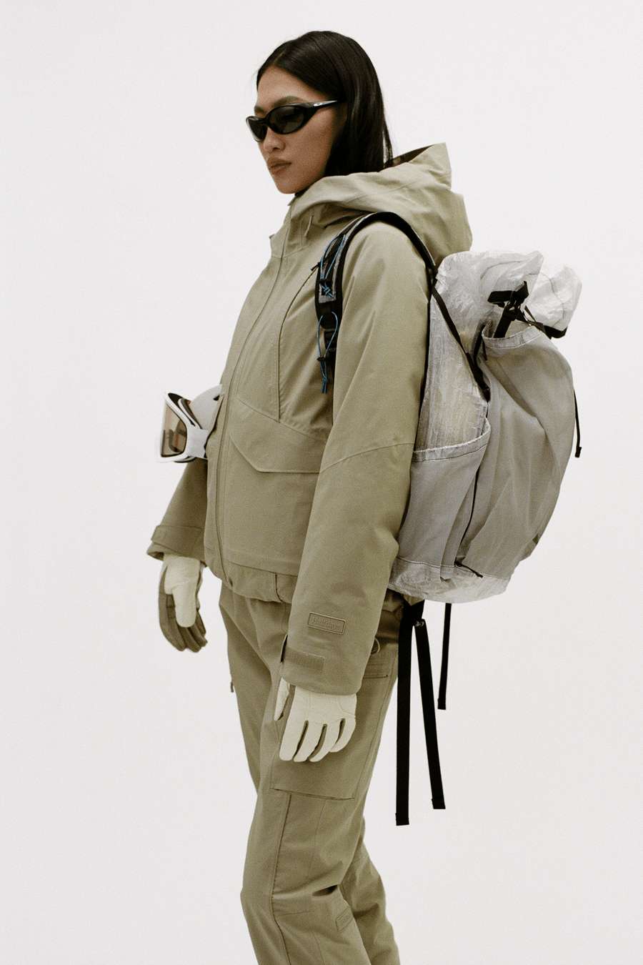 A model wearing a beige technical outdoor jacket and matching pants, accessorized with a large translucent gray backpack, white gloves, and black sunglasses, photographed against a plain light gray studio background.