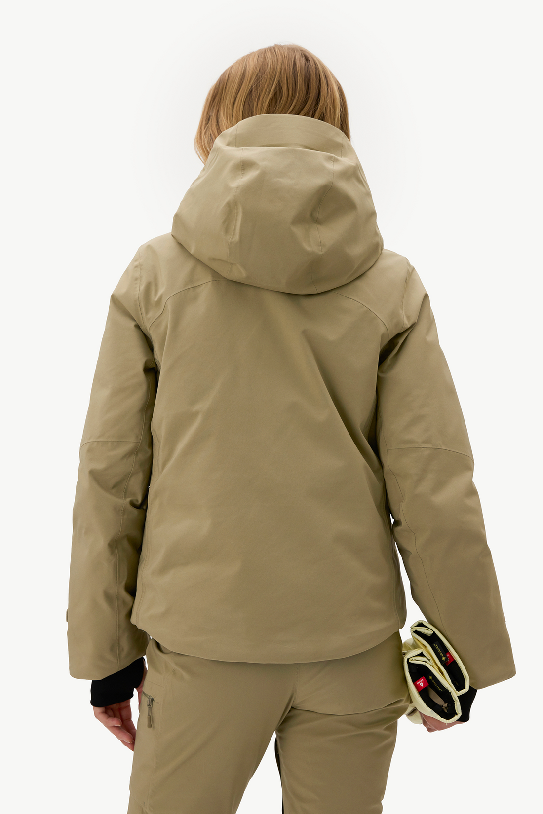 A person shown from the back wearing a hooded ski jacket and matching pants in a warm khaki-tan color, with a small black-and-yellow bag in one hand, against a clean white studio background.