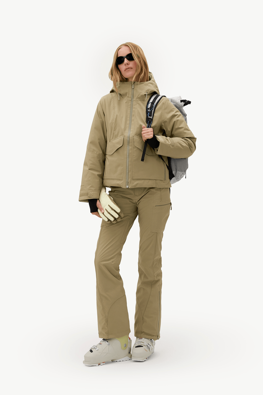 A woman modeling a beige ski jacket and matching ski pants, wearing a gray backpack, white gloves, and ski boots against a plain white studio background.