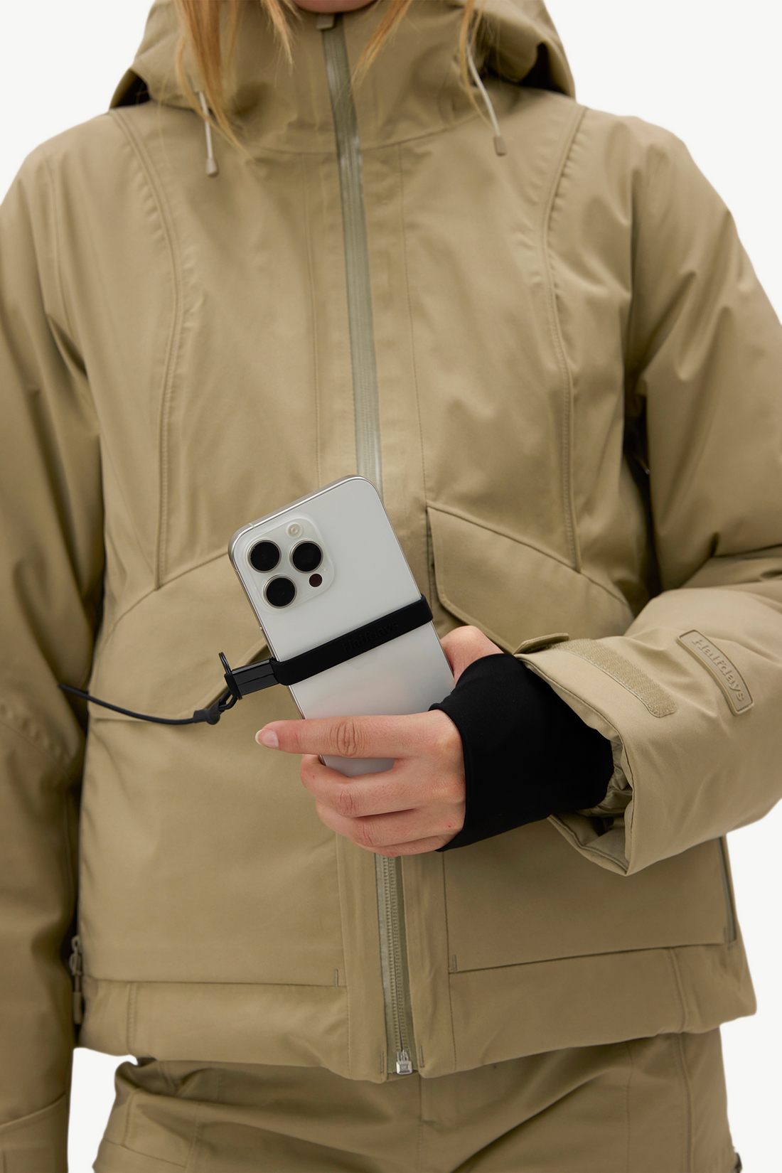 A close-up of a person wearing a beige hooded jacket and holding a white smartphone with a black band around it, against a plain light background.
