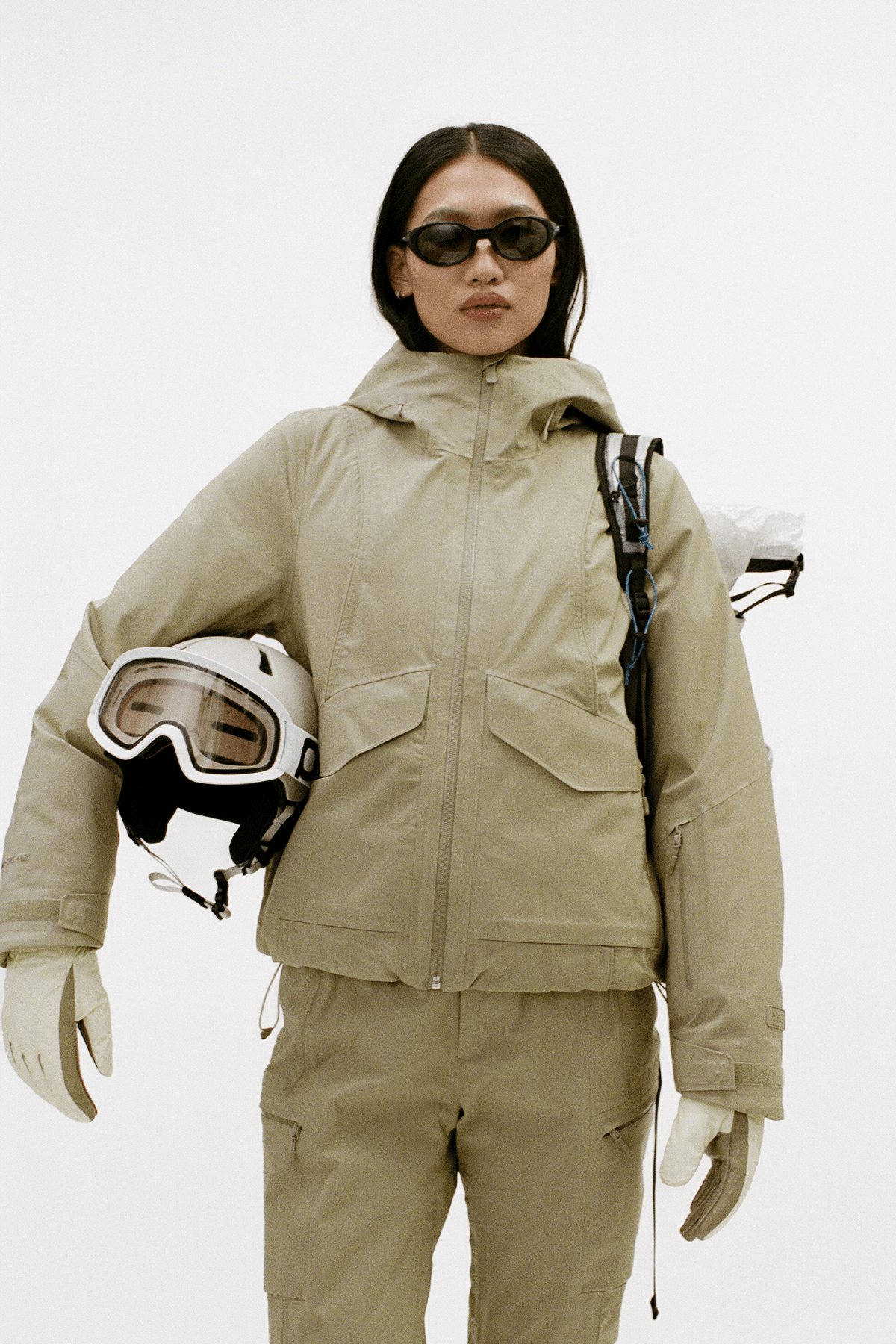 A model wearing a beige ski jacket and matching ski pants, holding a white ski helmet with goggles, set against a plain light gray studio background.