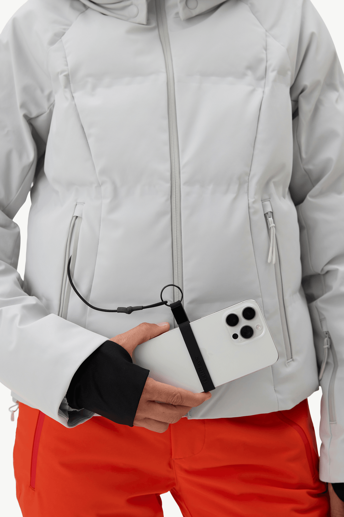 A close-up of a person wearing a light gray puffer jacket and bright orange ski pants, holding a white smartphone in their hand. A black strap with a small ring is attached near the zipper area of the jacket.