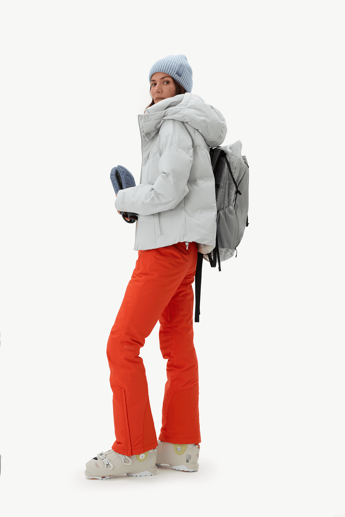 A woman wearing a light gray puffer jacket, bright orange ski pants, a light blue beanie, and a gray backpack, standing against a plain white studio background.