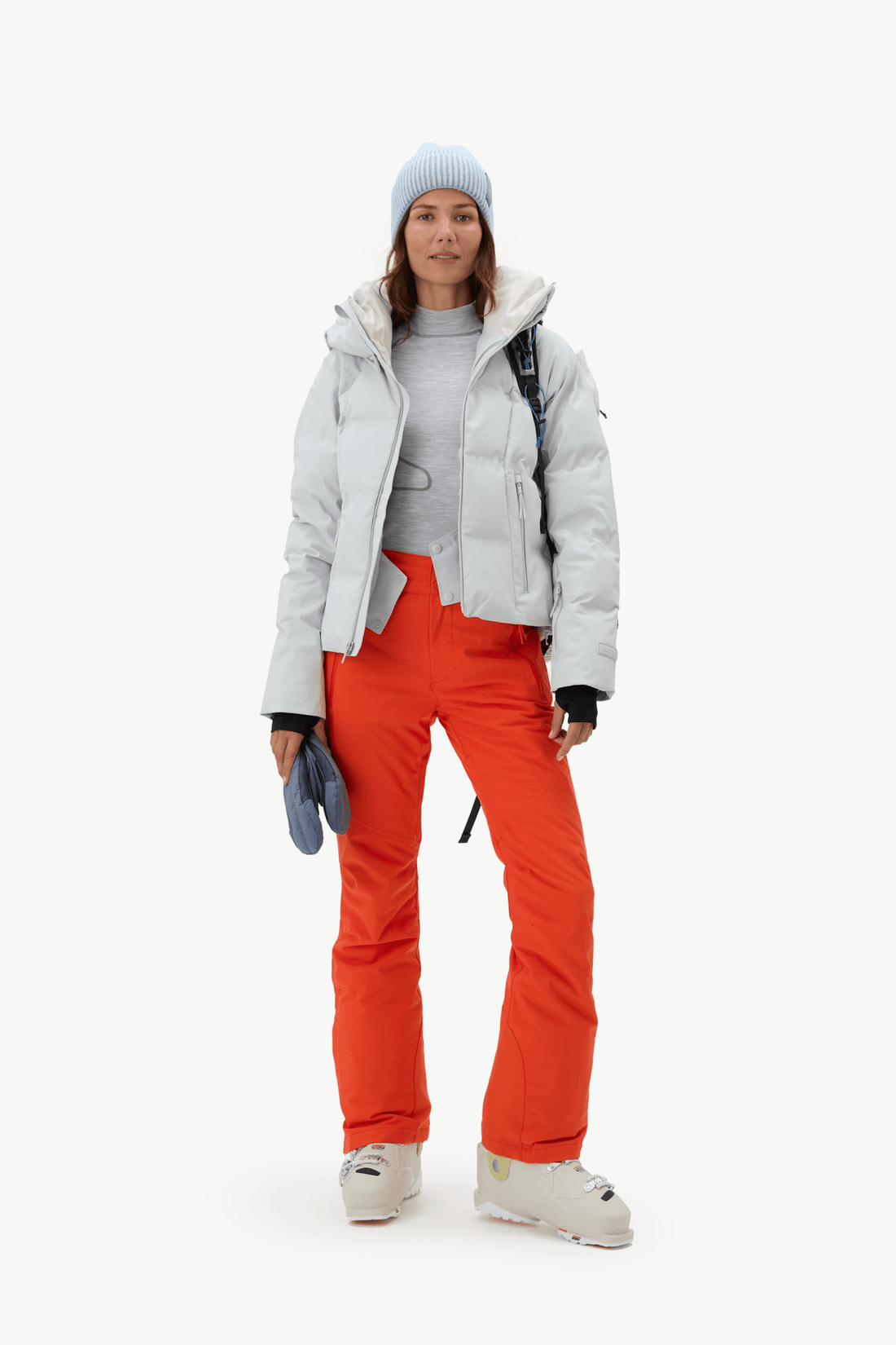 A model stands against a light gray studio background wearing a light gray puffer jacket, bright orange ski pants, a light blue knit beanie, a gray top, and white ski boots.
